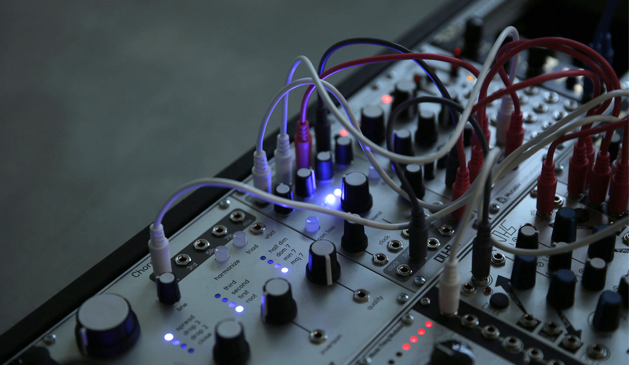 Modular synthesiser lies at an angle on a concrete floor, with a tangle of cables and flashing lights.